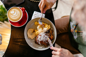 Top view of breakfast with scrambled eggs, croissant, coffee and sauce. Hands with fork and knife. Casual outfit, female hand with manicure, modern cafe lifestyle.