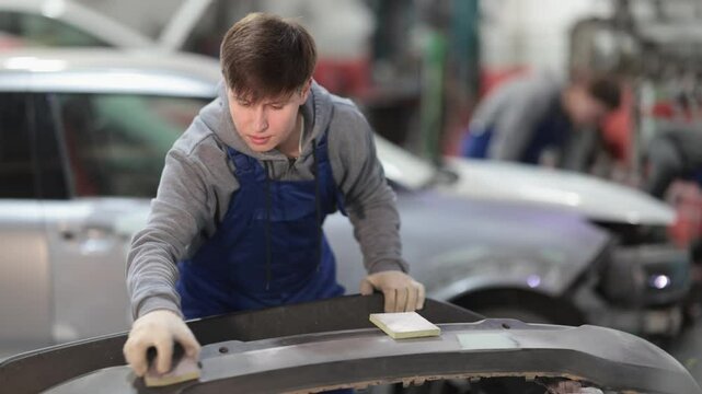 At service station, worker is engaged in straightening, eliminating deformations of car body. Auto mechanic at work. Guy auto mechanic handle polishes car bumper