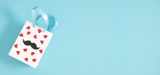 Fototapeta na wymiar Father's Day banner concept. Gift bag decorated with red confetti hearts and decorative mustaches on a light blue background. Flat lay.
