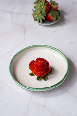 Strawberry carved into the shape of a rose lying on two leaves on a white and green ceramic plate on a marble table, next to a small bowl of strawberries