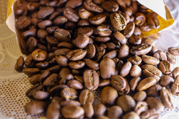 Fragrant roasted coffee beans are poured from an open kraft paper bag onto a white lace napkin. The...