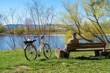 A man sits on a bench on the banks of the Rhine in Neuwied on a beautiful spring day. His bicycle is parked next to him on the grass