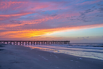 Sunrise over a wooden pier with waves breaking on the beach in the foreground.