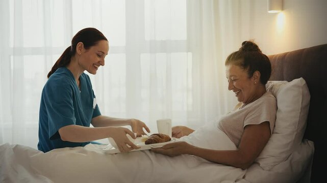 Retirement home worker bringing breakfast in bed to senior woman, caregiving