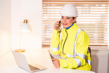 Female Construction site manager on call in bright safety jacket while working from office with laptop