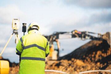 Site engineer surveying land with equipment while excavator operates in background