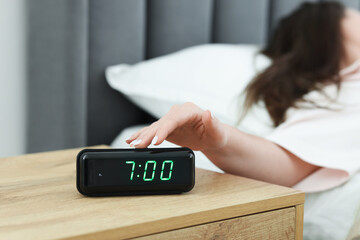 Woman turning off digital alarm clock on bedside table indoors, closeup