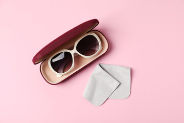 Case with stylish sunglasses and cleaning cloth on light pink background, top view