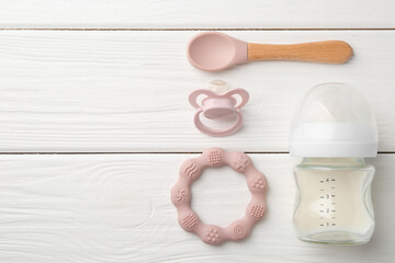 Feeding bottle with milk, spoon, pacifier and teether on white wooden table, flat lay. Space for...