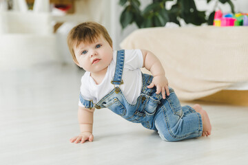 cute baby in denim overalls learning to crawl on the floor in a bright room, child smiling,...