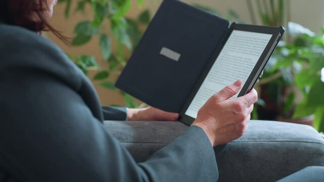 Middle-aged woman reading digital book on e-reader while relaxing on gray couch surrounded by indoor plants in cozy living room environment during peaceful afternoon moment of solitude