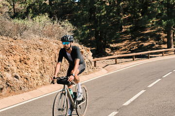 A cyclist in black cycling apparel rides a gravel bike along a scenic mountain road. The sun shines brightly, highlighting the stunning natural landscape.