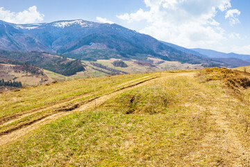 Fototapeta premium rural road through the hill with grassy meadow. sunny day. carpathian mountain ridge in the distance. alpine countryside of ukraine in early spring. fluffy clouds on the blue sky