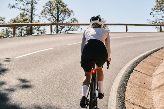 Rear view of a female cyclist riding a road bike on a scenic mountain road.  Sunlight illuminates the scene, showcasing the cyclist's athletic form and the beautiful landscape.