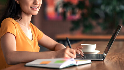 Young business woman planning her day, writing in notepad, sitting in front of laptop, drinking...