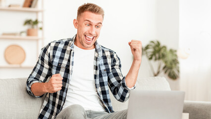 Fototapeta premium Yeah I Did It. Young man with raised arms working on laptop and celebrating success, sitting on the couch at home