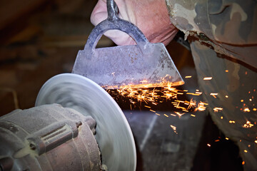A man's hand sharpens a hoe on an electric sharpening machine in the village. Sparks from the...