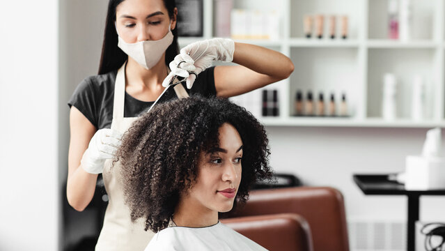 Hairdresser in protective mask cutting hair of curly african american client in beauty salon, free space