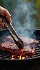 Flap steak being flipped on outdoor grill with kitchen tongs, blue-gray smoke rising as juices hit coals, flames leaping around edges, showing sear marks forming