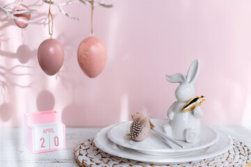 Table place setting with easter bunny figure, eggs, empty glass and wooden Perpetual calendar indicating March 31 on pastel pink background. Happy easter greeting or invitation card