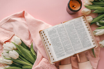 Open Bible held in hands over a pink background, surrounded by white tulips, a lit candle, and a...