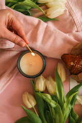 Close-up of a hand lighting a candle with a match. Soft focus on white tulips and a pink jacket in...
