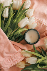 Cozy still life with a burnt candle in an amber glass jar surrounded by white tulips, a pink denim...