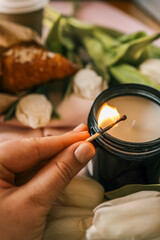 Close-up of a hand lighting a candle with a match. Soft focus on white tulips and a pink jacket in...