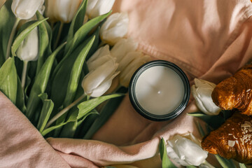 Cozy still life with a burnt candle in an amber glass jar surrounded by white tulips, a pink denim...