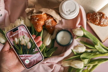 Smartphone in hand displaying "Women's Day" call screen, surrounded by white tulips, croissants, a candle, and takeaway coffee on a soft pink background.