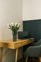 Minimalist workspace with a wooden desk, laptop, and white tulips in a glass vase. Cozy and modern interior with soft lighting and a gray chair on a furry rug