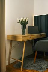 Minimalist workspace with a wooden desk, laptop, and white tulips in a glass vase. Cozy and modern interior with soft lighting and a gray chair on a furry rug