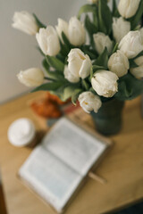 Close-up of a bouquet of white tulips in a vase on a wooden table with an open Bible and a coffee...