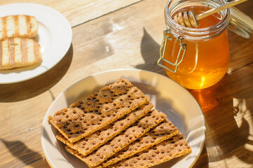 A jar of honey sits beside crispy bread on a large plate, resting on a rustic wooden table. Soft...
