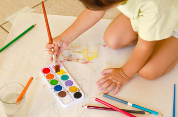 A little girl painting with watercolors and a brush on a paper on the floor. 