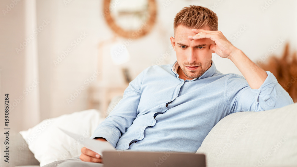 Poster foused man working from home, sitting on sofa, holding document and looking at personal computer