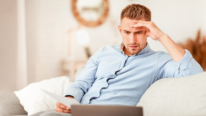 Foused man working from home, sitting on sofa, holding document and looking at personal computer