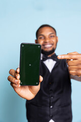African american bellhop presenting green screen on a phone, posing with hospitality industry suit and tie in studio. Young hotelier personnel creates an isolated advertisement.