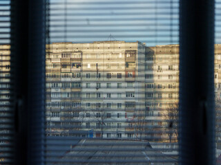 Sunlight filters through closed window blinds, illuminating a residential building across the street. The structure features multiple balconies and windows, showcasing urban life in a city.