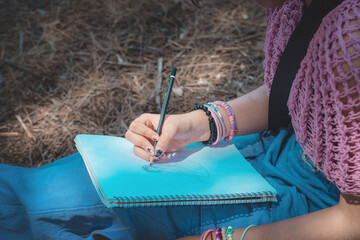 Young woman is drawing in her notebook while sitting in the forest.