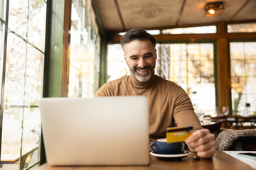 A man smiles while holding a credit card and using his laptop for online shopping in a cozy café setting.