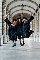 Three cheerful students wearing graduation gowns and caps jumping together, celebrating their...
