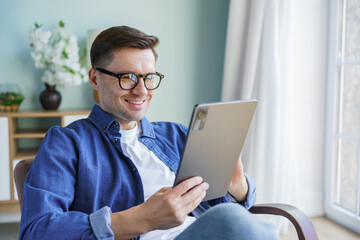 Smiling Man Enjoys Using a Tablet While Sitting in a Cozy Living Room on a Sunny Day
