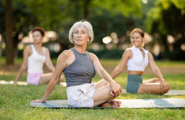 Sedulous old lady and group of women doing spinal twist pose of yoga on mat in modern light city...