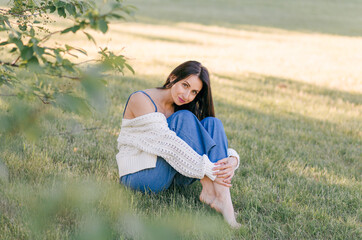 A young beautiful woman is sitting on the green grass, outdoor on a sunny morning. Relax in nature, happiness. Enjoying Summer days outdoors