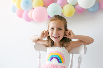 Portrait of little happy girl in dress, sitting on the chair with colorful balloons on a white background, studio shoot. Celebrations, birthday and playing concept.