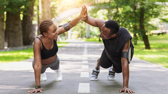 Motivated black couple giving high-five to each other while working out outdoors, standing in plank or doing push ups in park, clapping hands