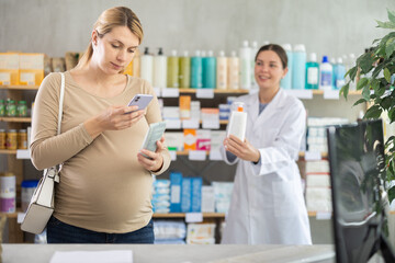Fototapeta premium Pregnant woman in pharmacy scans QR code on antifungal cream product, checks certificate on manufacturers website. Girl pharmacist offer similar product in background.