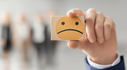 Businessman holding a wooden block with a sad face emoji, symbolizing dissatisfaction or negative feedback, with blurred colleagues in the background.