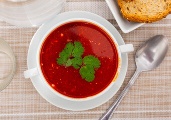 Appetizing beetroot soup with beef decorated by greens served in bowl with cutlery and bread in restaurant